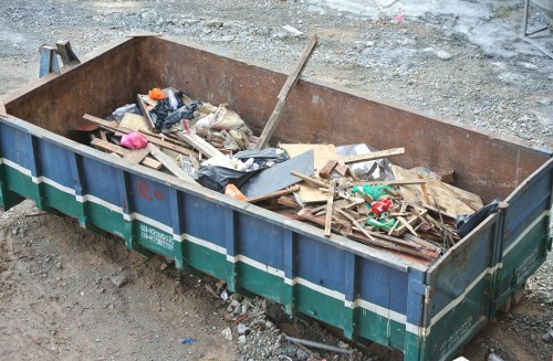 A large, rectangular metal skip bin positioned outdoors on a gravel surface, filled with assorted construction and renovation waste including wooden planks, broken panels, black plastic bags, and various discarded materials. The skip has a rusted, weathered appearance and is situated in an open area likely within a commercial or industrial site. Surrounding the bin are rough, uneven ground with loose stones and dirt, indicating a work zone or site for rubbish removal services. The overall scene reflects a typical disposal container used for clearing waste from building projects, with no ongoing activity visible. The presence of Commercial Waste Hillingdon’s service suggests a local rubbish removal solution in the Hillingdon area, suitable for handling diverse waste streams efficiently.