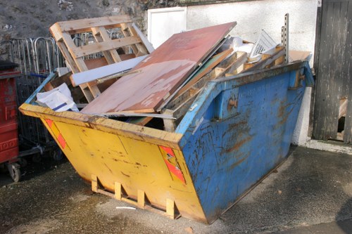 A large, metal skip positioned on a paved outdoor surface, painted in blue with visible signs of rust and wear along its edges. The skip is filled with various construction and renovation debris, including wooden pallets, particle board, painted MDF panels, and other miscellaneous waste materials. Some wood pieces are leaning outward, protruding over the top edge, while others are stacked haphazardly inside. The debris varies in colour and texture, with some items showing painted surfaces and others untreated wood. The environment appears to be an outdoor area adjacent to a building, with a white wall and a weathered black wooden fence in the background. The overall scene suggests a typical waste removal scenario managed by Commercial Waste Hillingdon, serving local businesses and construction sites in the area for rubbish clearance and disposal.