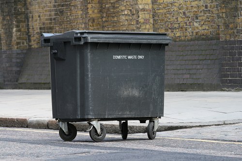 A large, dark grey plastic rubbish bin with a hinged lid, positioned on a paved sidewalk next to a curb and street. The bin has four small black wheels, with a sticker on the front indicating it is for 'domestic waste only.' The background features a brick wall with weathered yellowish bricks and a concrete retaining wall, suggesting an urban or suburban setting in Hillingdon. The surface of the bin appears to have some dirt or smudges, and it is placed upright on a slight incline from the main road. The scene is captured during daylight hours, with natural light illuminating the area. This image, associated with Commercial Waste Hillingdon, illustrates the typical waste container used for local rubbish collection services in the area, providing a clear visual reference for waste management and removal in Hillingdon.