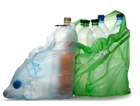 A collection of used plastic bottles of various sizes and colours, including clear, blue, and green, are gathered together inside a transparent plastic bag with a light blue tint, partially folded over at the top. The bottles, some with visible caps and labels, are arranged in a haphazard manner, with the green bottles leaning to the right and the clear ones on the left. The plastic bag appears stretched and crinkled, sitting on a plain white surface with no background visible. This image illustrates typical waste packaging ready for disposal or collection, relevant to rubbish removal services such as those offered by Commercial Waste Hillingdon in the local area, possibly involving the collection of recyclable plastics from commercial or residential settings in Hillingdon and surrounding postcodes.