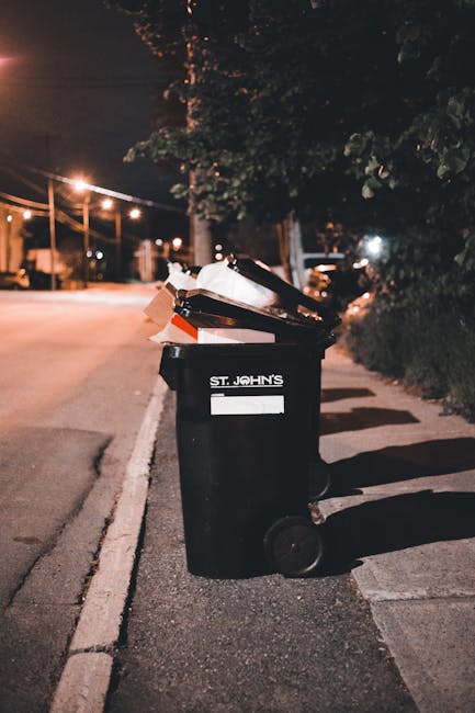 An outdoor scene showing a large collection of mixed waste and rubbish overflowing from multiple trash bins placed on a paved sidewalk next to a metal railing. The waste includes crumpled cardboard bo